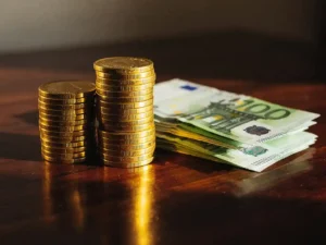 Stapel gouden munten naast 100-euro bankbiljetten op donkere houten tafel met warm zonlicht en zachte schaduwen